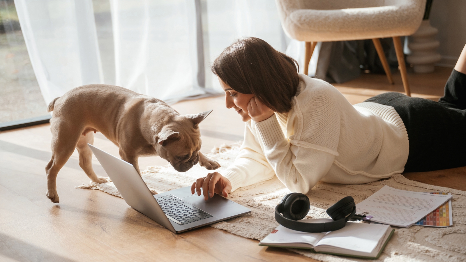 A woman lies on a rug using a laptop while her French Bulldog stands next to it, peering closely at the screen and placing its paw on her hand.